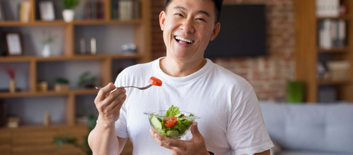 Happy asian mature man holding fork and bowl with fresh vegetable salad, eating healthy lunch after domestic training. Active middle aged male having balanced meal