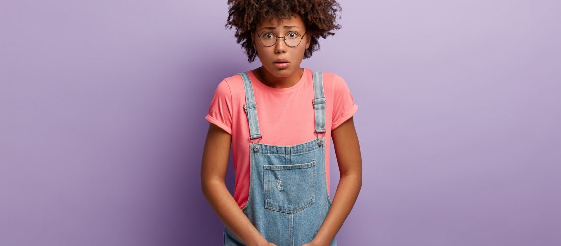 Dissatisfied girl keeps hands on crotch, presses lower abdomen, needs toilet badly, has syndrome of cystitis, wears spectacles, pink t shirt and denim sarafan, isolated on purple wall. Health problem